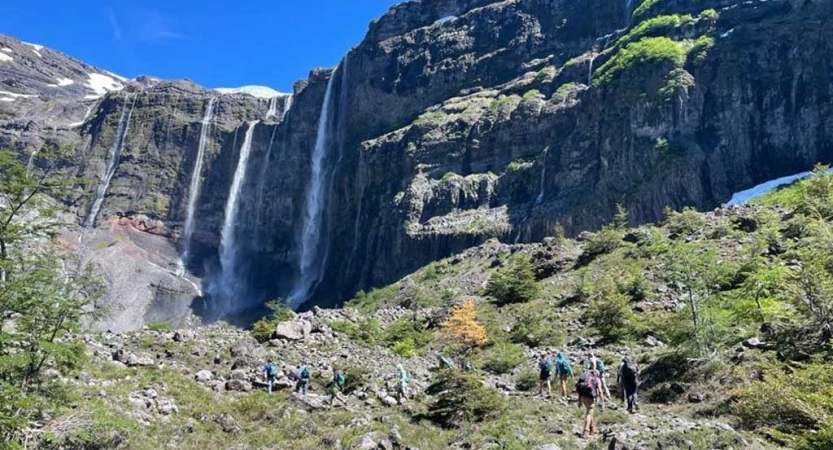 Several waterfalls stream over a tall cliff. In the foreground, a group of hikers appear very small making their way toward the falls. 